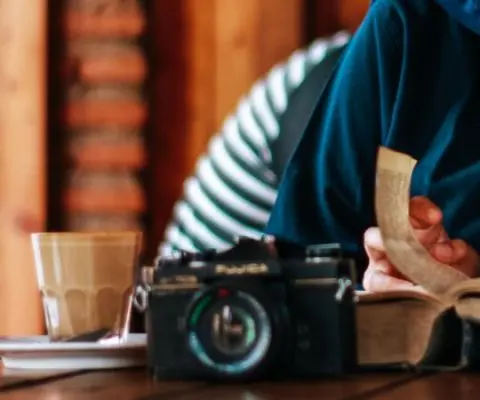 person sitting at table with camera and drink reading book