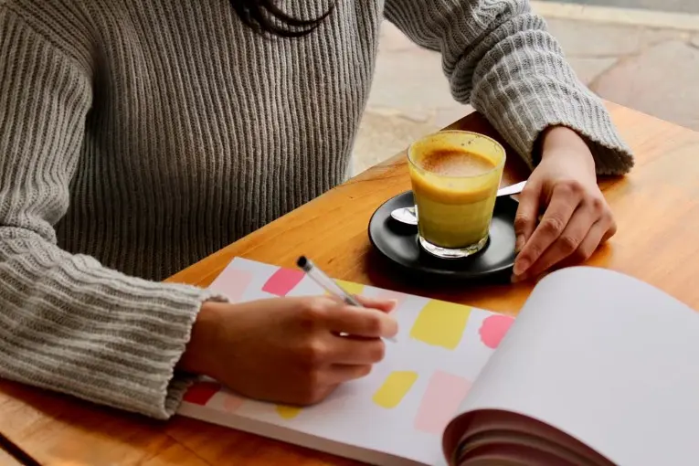 person writing on a notebook with a coffee at the side
