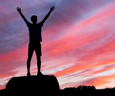 Silhouette of person standing on rocks with arms outstretched against pink sunset