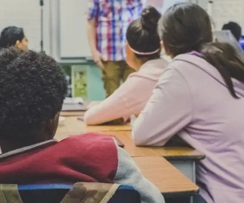 young students sitting around table looking at person standing at the front of classroom