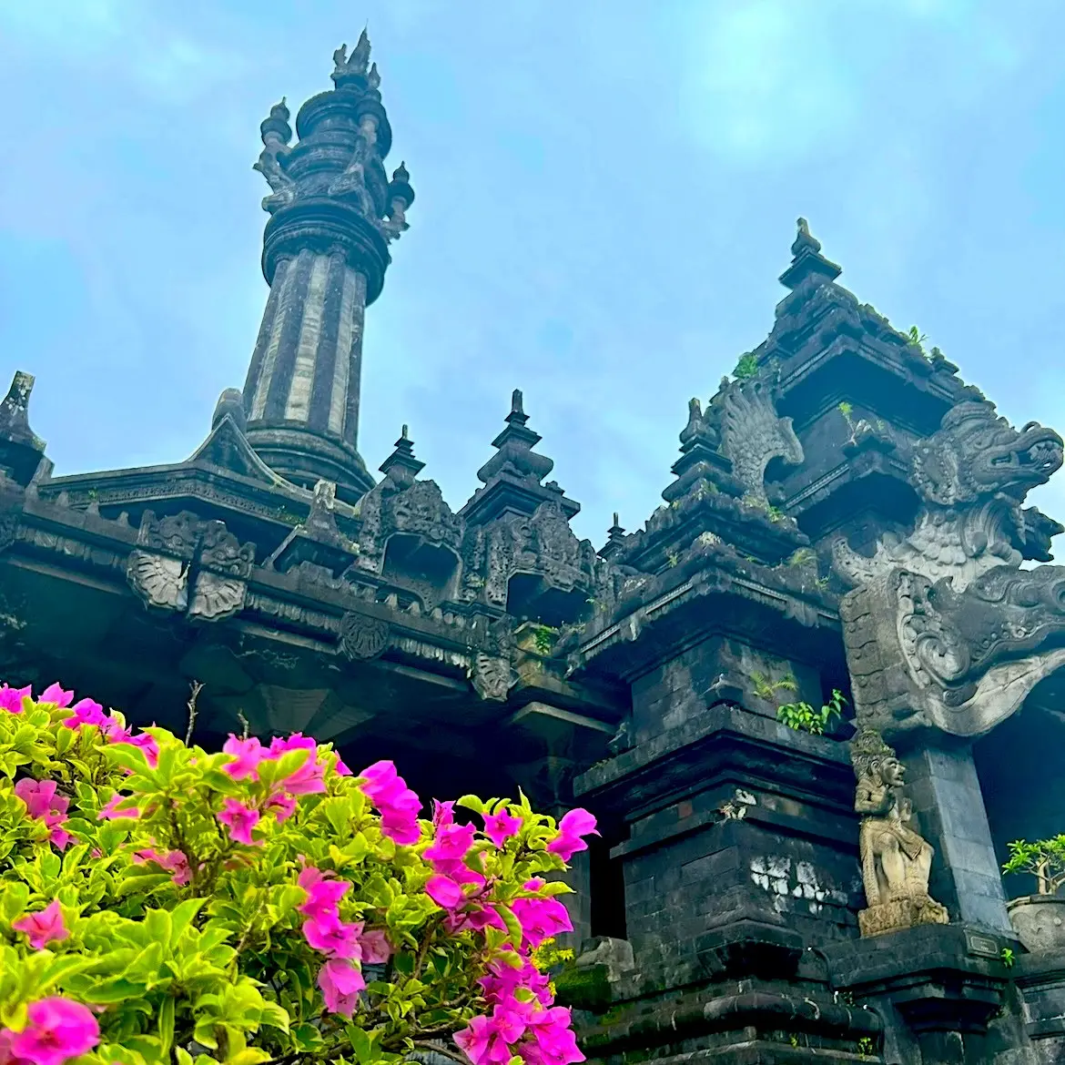 Ornate temple with intricate carvings and a tall spire against a cloudy sky, accented by bright pink flowers in the foreground, creating a serene ambiance.
